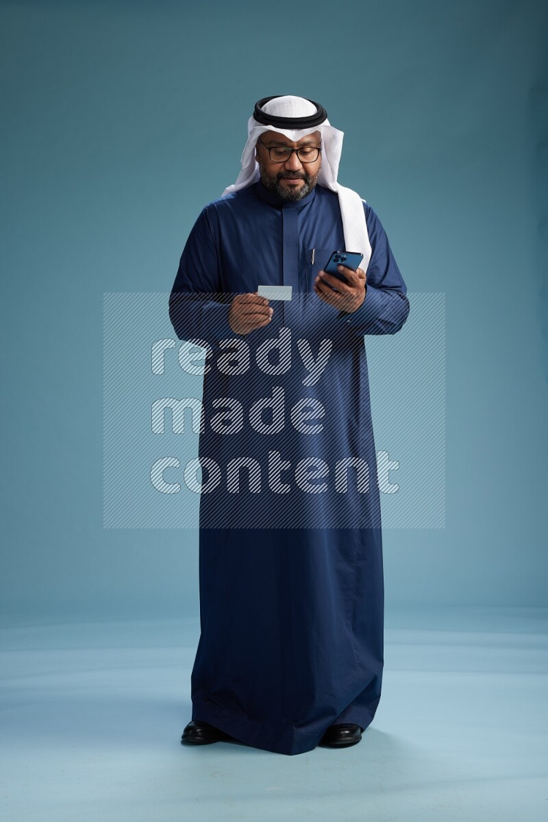 Saudi Man with shimag Standing holding ATM card while talking on phone on blue background