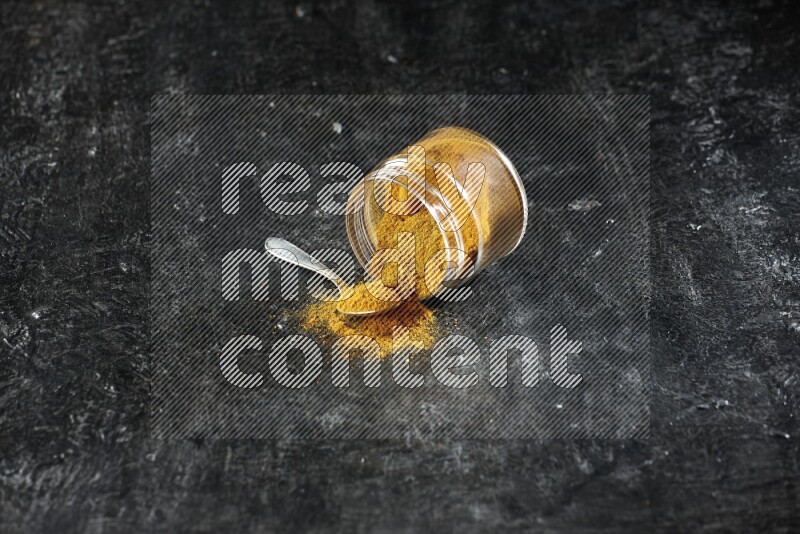 وعاء زجاجي مقلوب وملعقة معدنية ممتلئان ببودرة الكركم والبودرة تناثرت منه على خلفية سوداء