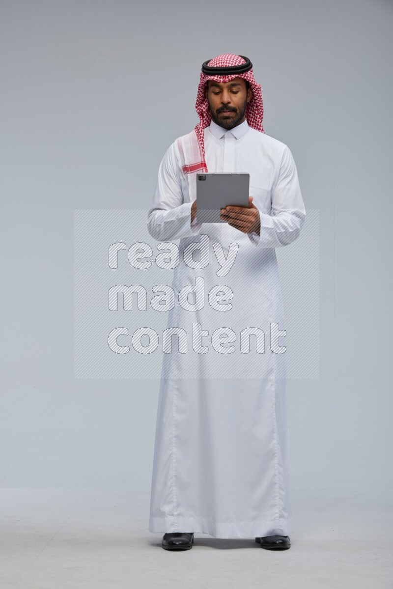 Saudi man Wearing Thob and shomag standing working on tablet on Gray background