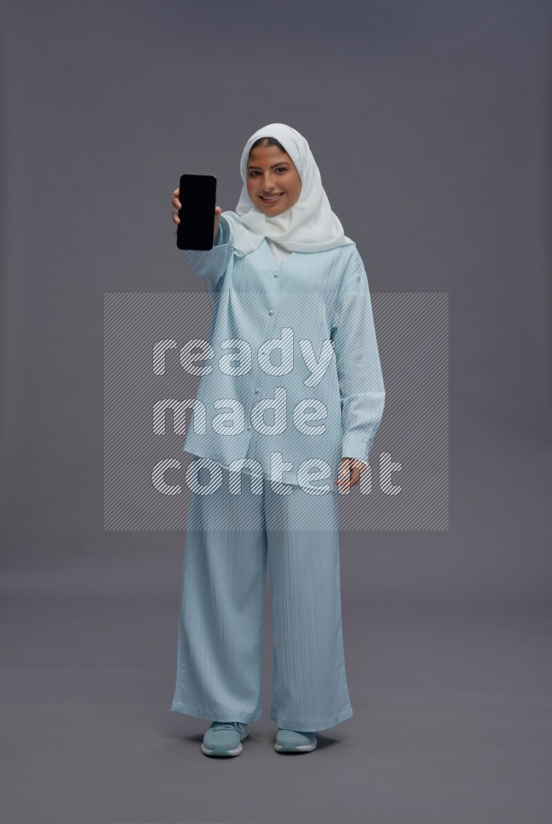 Saudi woman wearing hijab clothes standing showing phone to camera on gray background