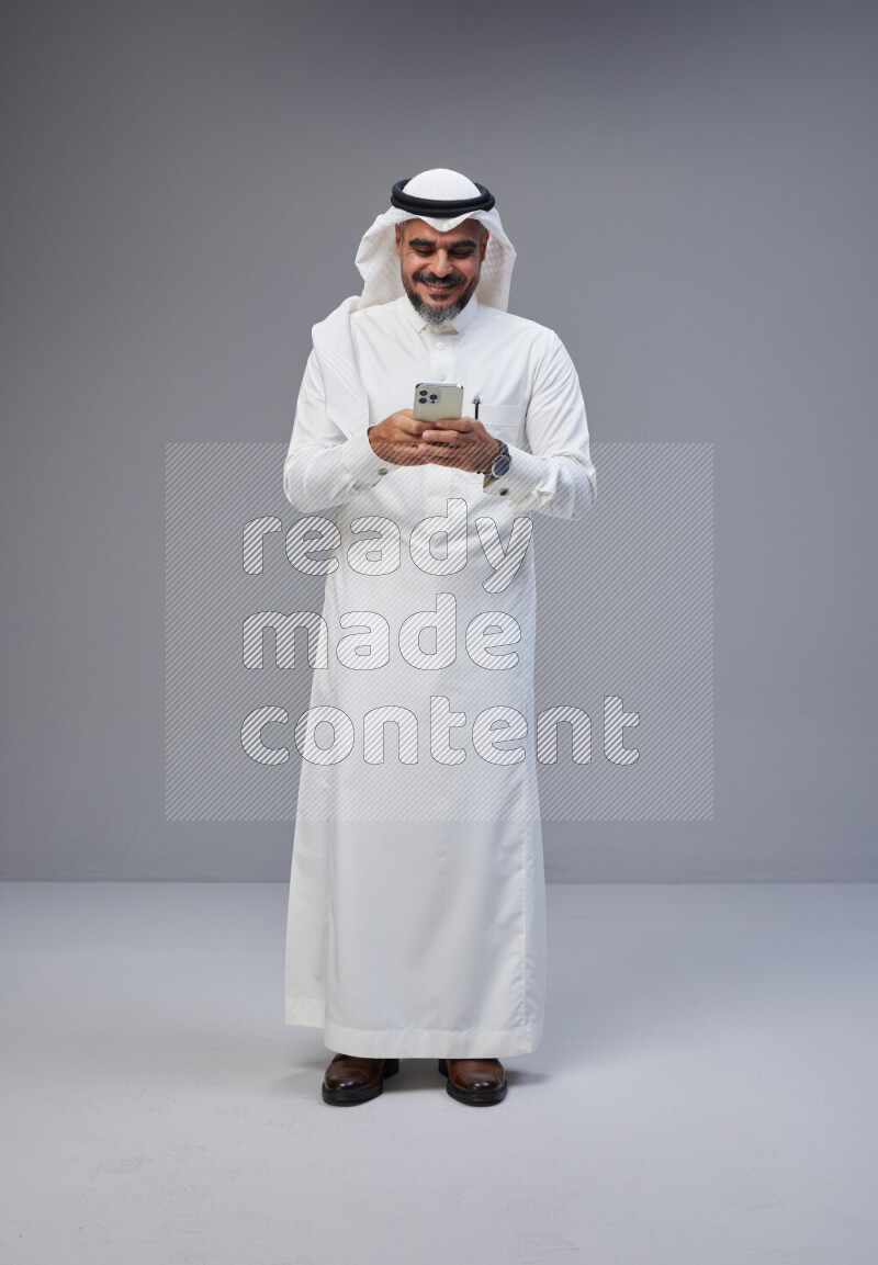 Saudi man Wearing Thob and white Shomag standing texting on phone on Gray background
