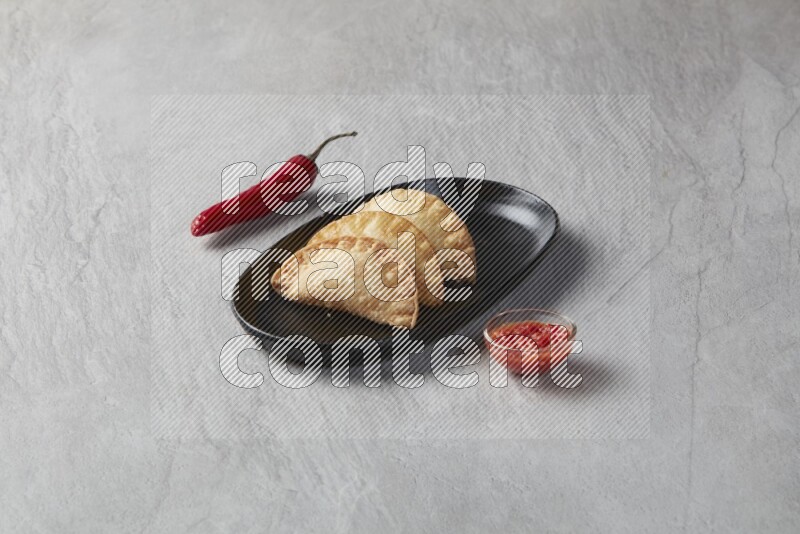 Three fried sambosas in an oval shaped black plate and a red sauce in a black round ramekin on a gray background