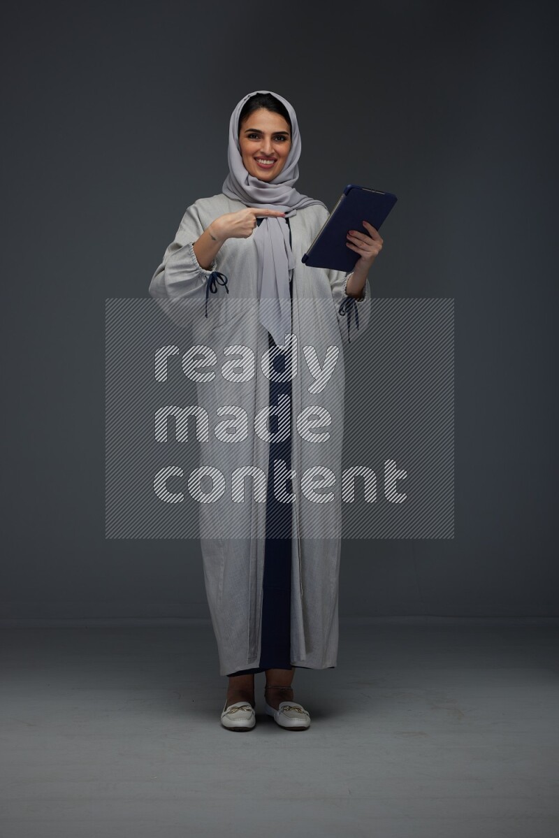 A Saudi woman wearing a light gray Abaya and head scarf standing holding a phone and pointing with the other hand eye level on a grey background