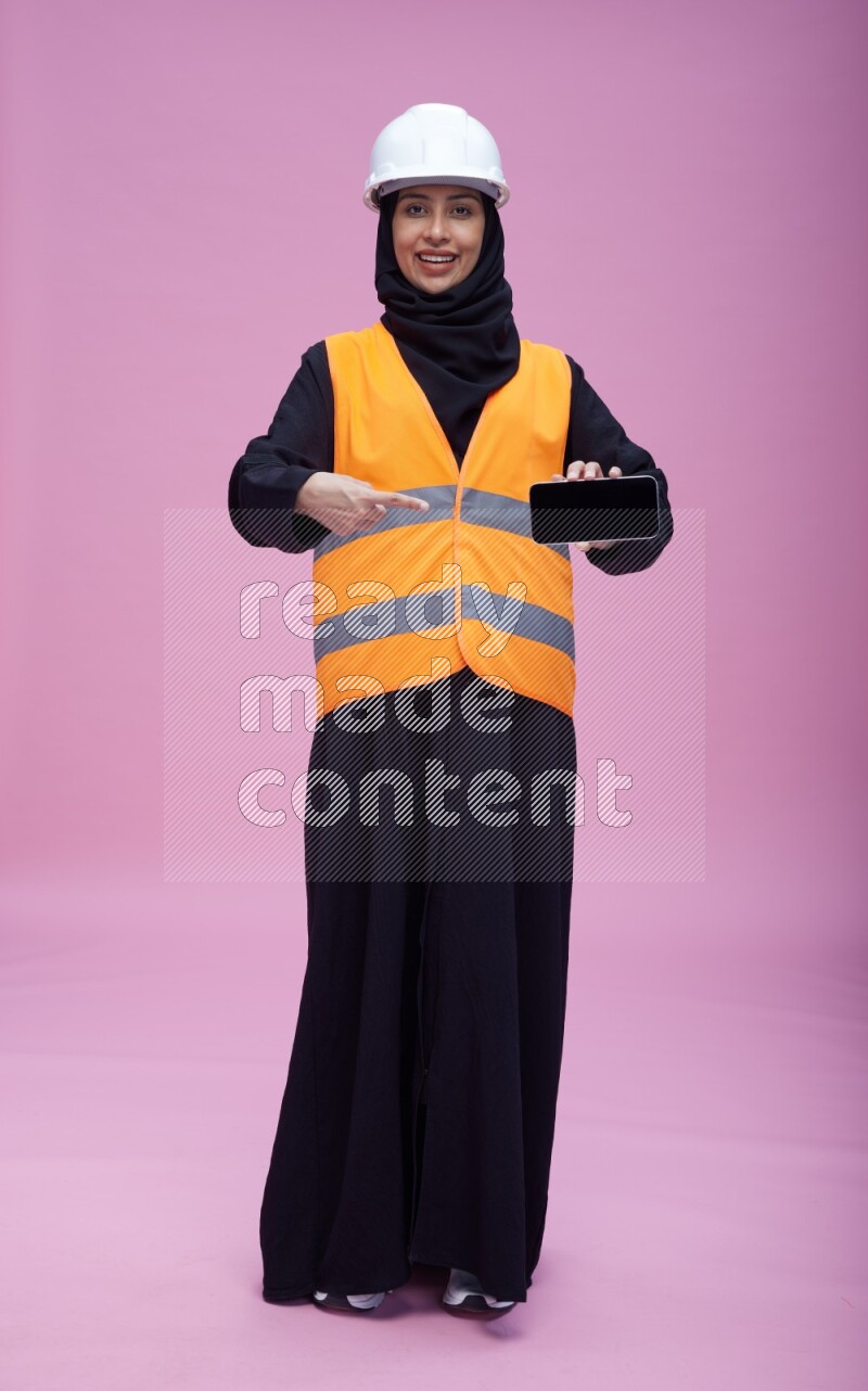 Saudi woman wearing Abaya with engineer vest and helmet standing showing phone to camera on pink background