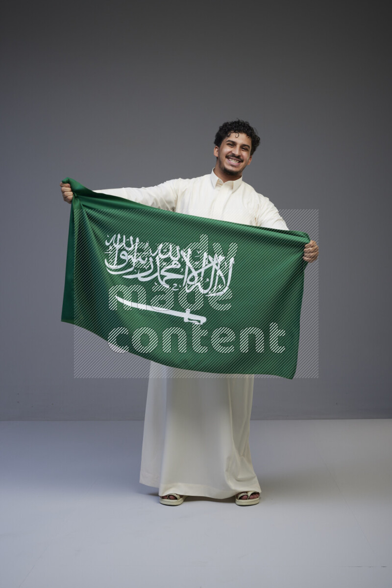 A Saudi man standing wearing thob holding big Saudi flag on gray background