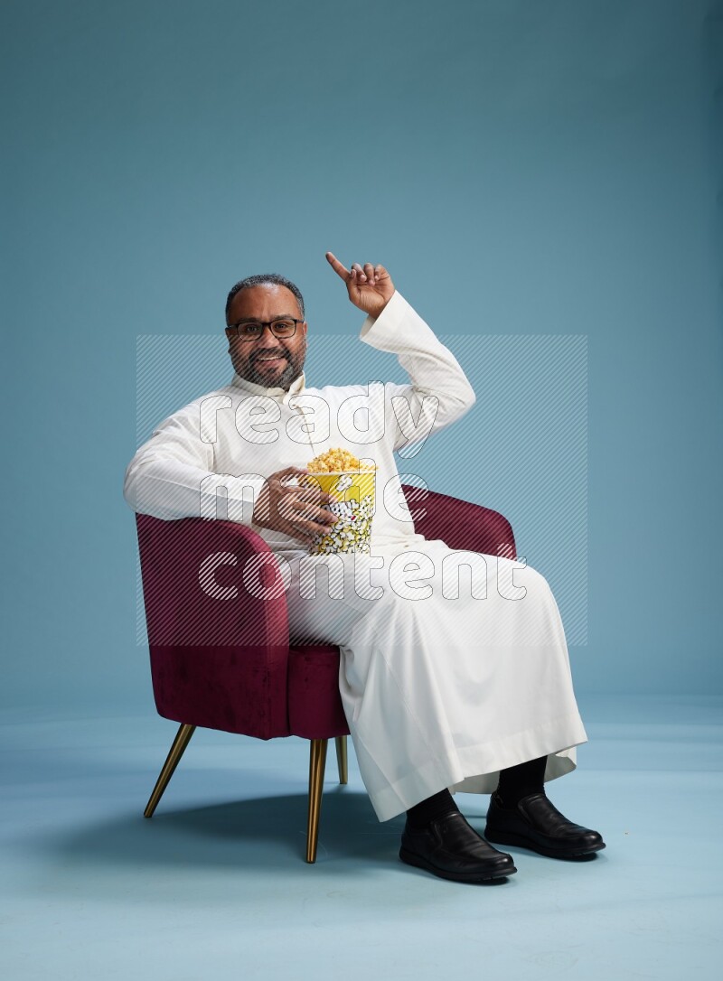 Saudi Man without shimag sitting on chair eating popcorn on blue background
