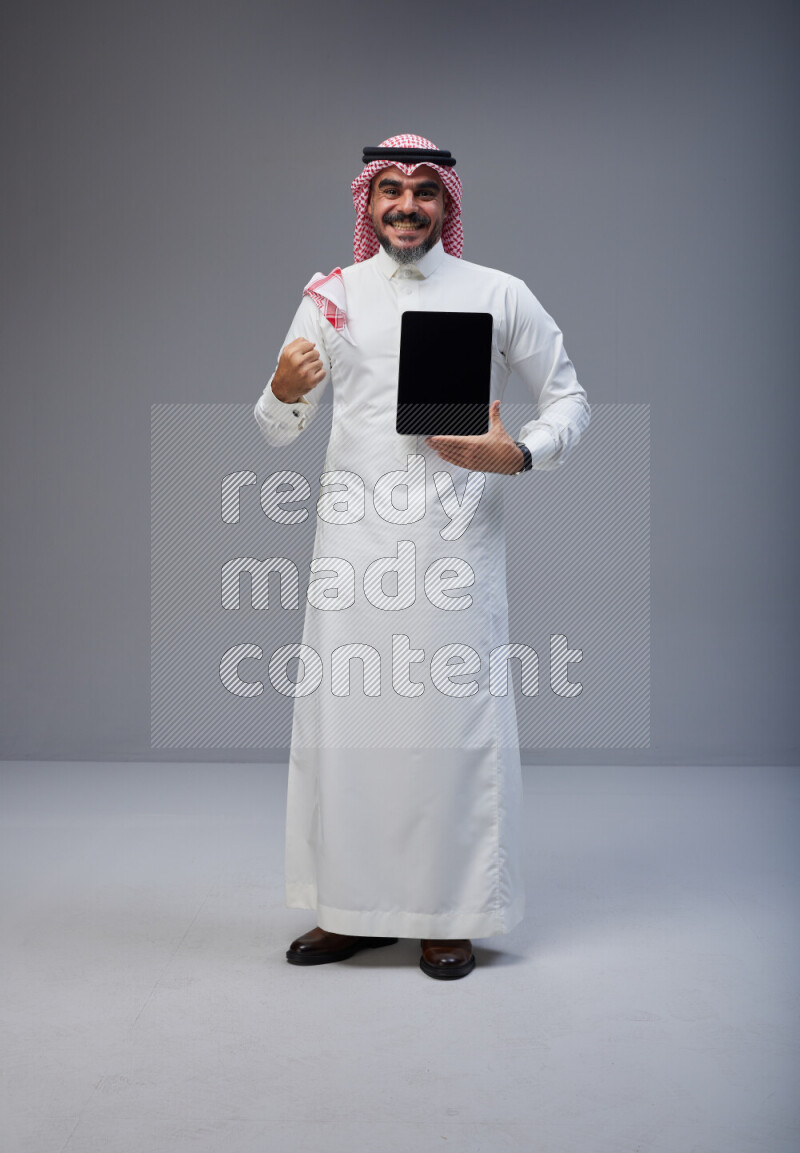 Saudi man Wearing Thob and red Shomag standing showing tablet to camera on Gray background