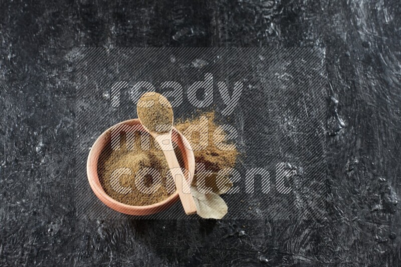 A wooden bowl and spoon full of cumin powder on a textured black flooring