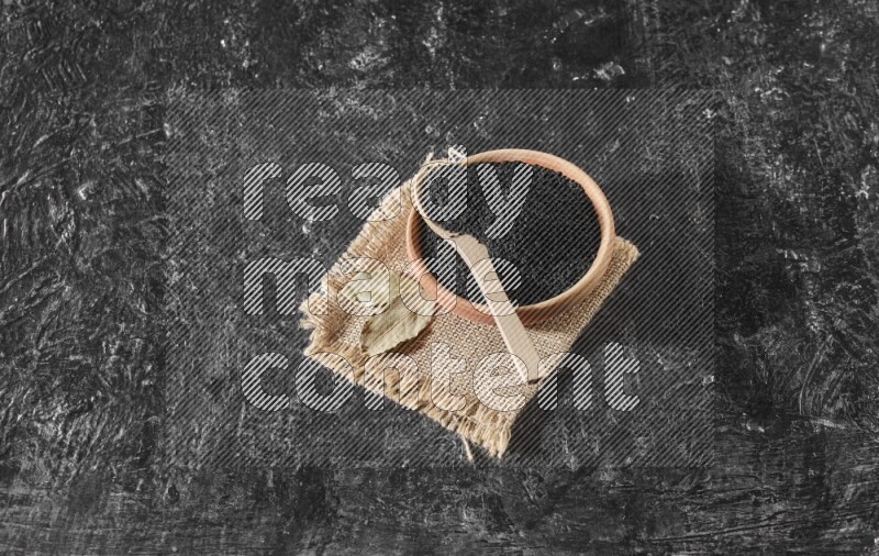 وعاء خشبي مع ملعقة خشبية ممتلئان ببذور حبة البركة علي قطعة من القماش علي خلفية سوداء