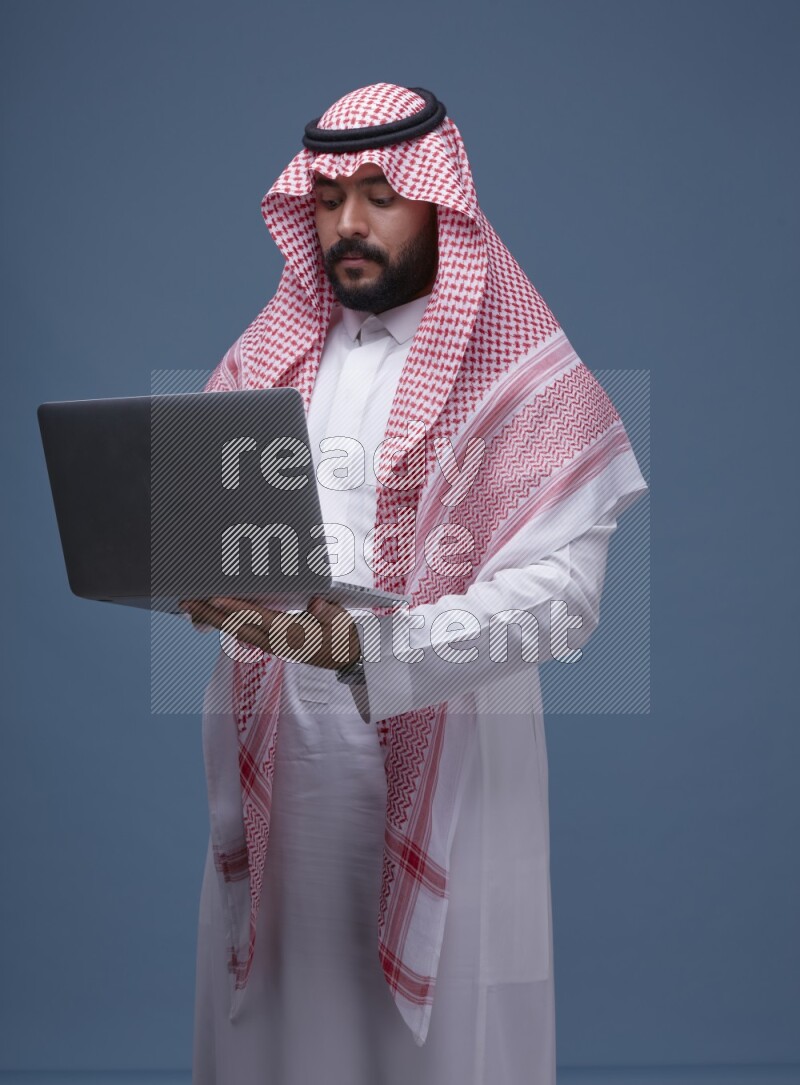 A man standing  with a laptop on Blue Background wearing Saudi Thob and Shomag