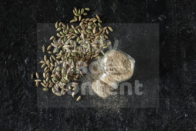 وعاء زجاجي للتوابل مقلوب وملعقة معدنية ممتلئة ببودرة الهيل وحبوب الهيل متناثرة على خلفية سوداء