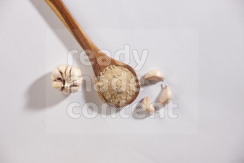 A wooden ladle full of garlic powder with a whole garlic bulb and some cloves beside it on a white flooring