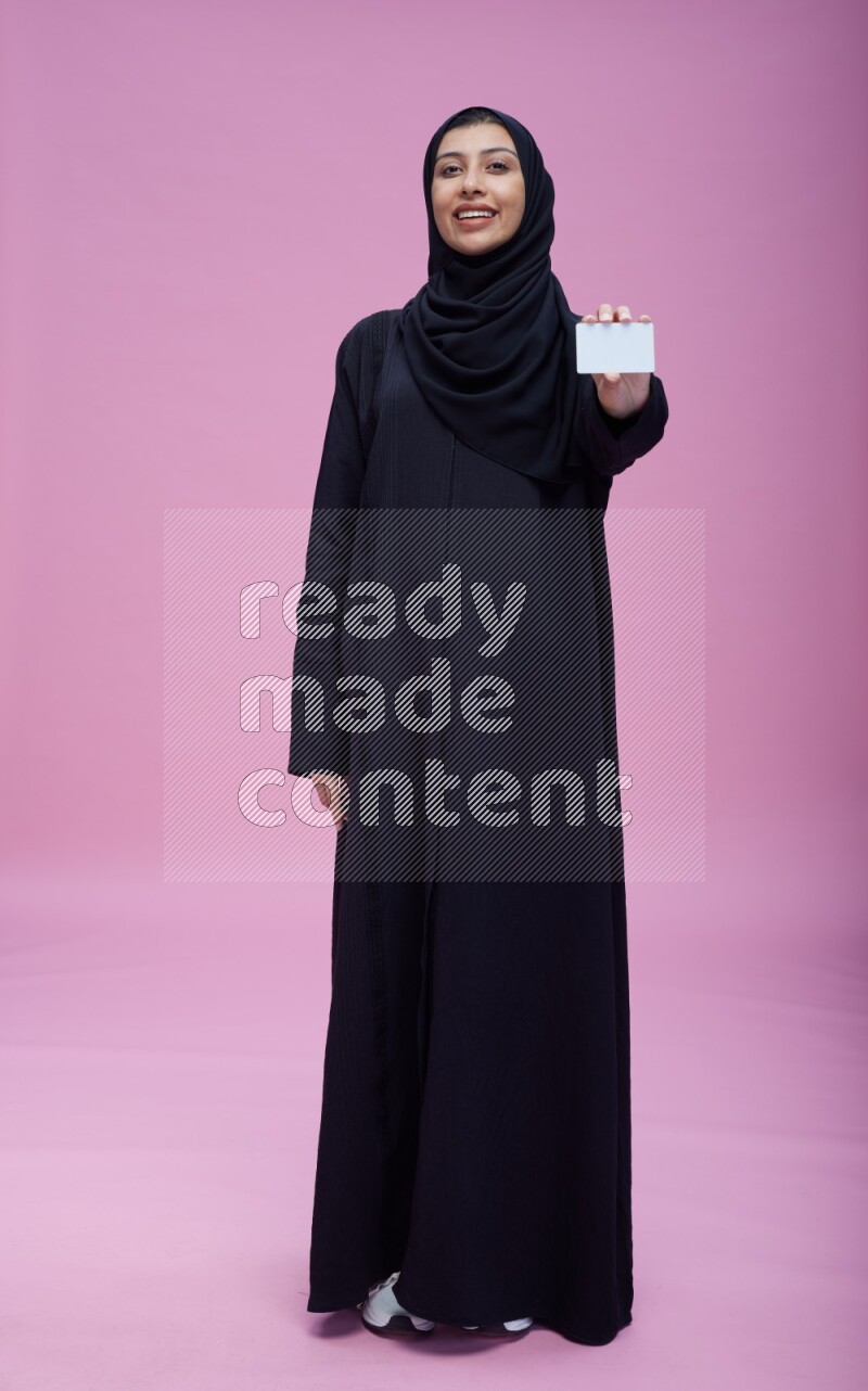 Saudi woman wearing Abaya standing holding ATM card on pink background