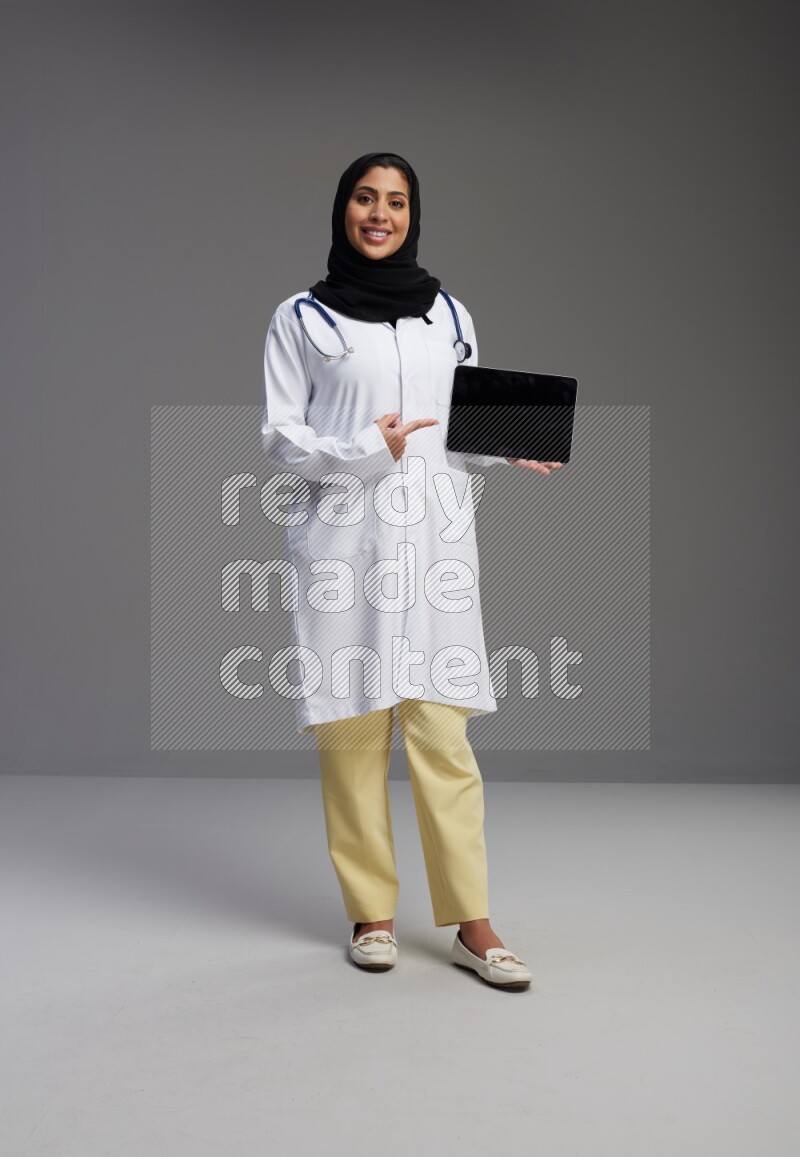 Saudi woman wearing lab coat with stethoscope standing showing tablet to camera with sign in the back on Gray background