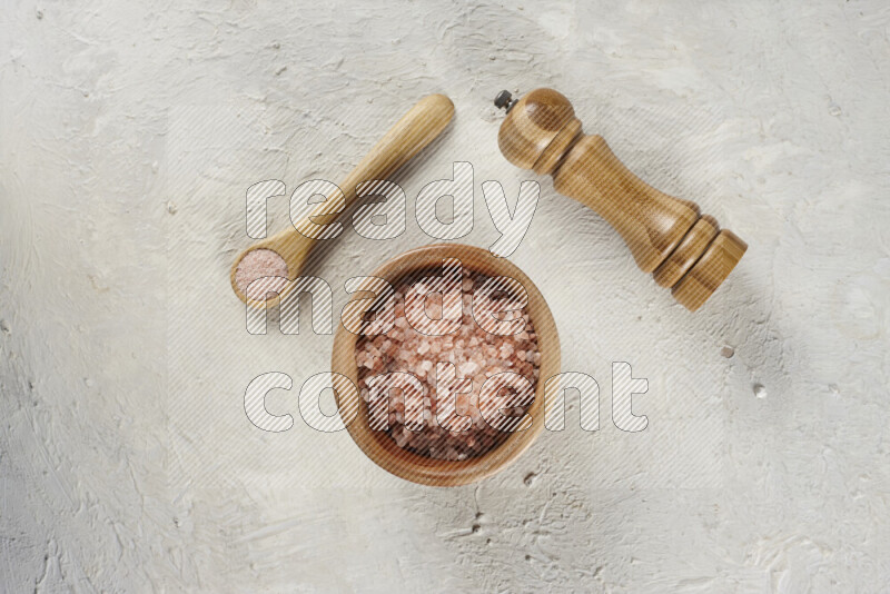 A wooden bowl and spoon filled with coarse pink himalayan salt and a wooden grinder beside them on white background