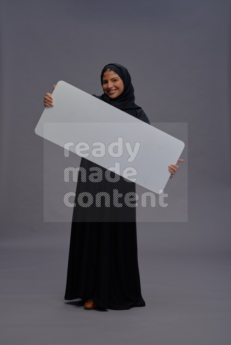 Saudi woman wearing Abaya standing holding white board on gray background
