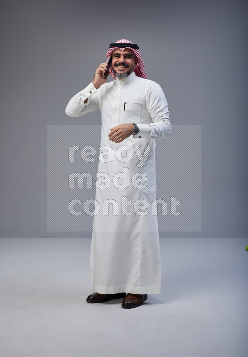 Saudi man Wearing Thob and red Shomag standing talking on phone on Gray background