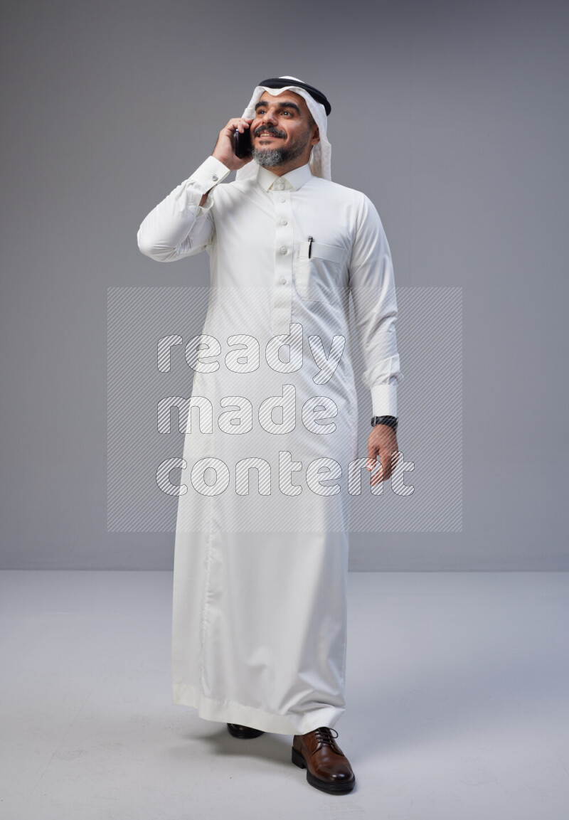 Saudi man Wearing Thob and white Shomag standing talking on phone on Gray background