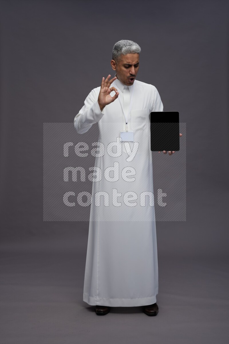 Saudi man wearing thob with neck strap employee badge standing showing tablet to camera on gray background