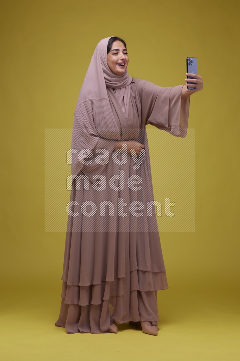 A woman taking a Selfie on a Yellow Background wearing Brown Abaya with Hijab