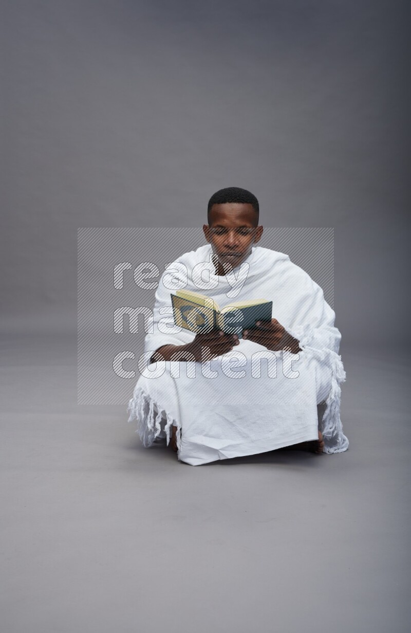 A man wearing Ehram sitting on floor reading quran on gray background