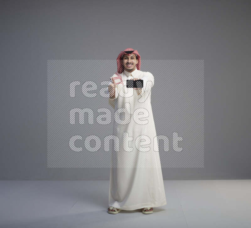 A Saudi man standing wearing thob and red shomag showing phone to camera on gray background