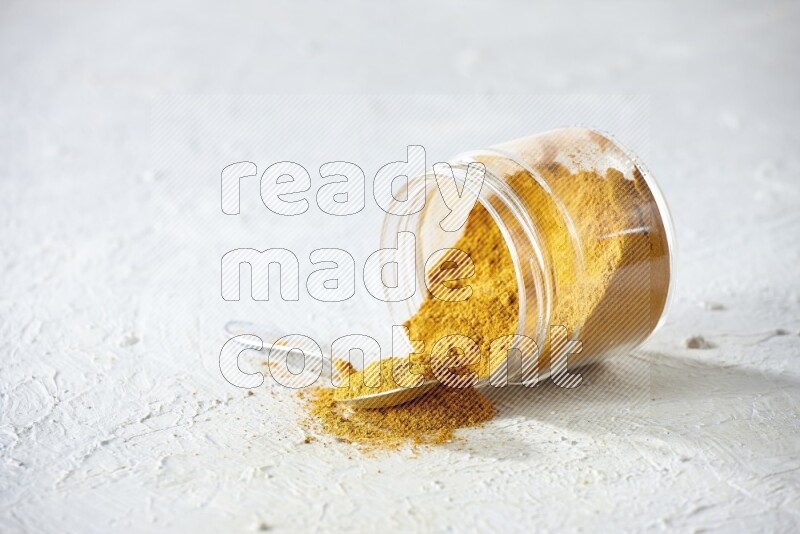 A flipped glass spice jar and a metal spoon full of turmeric powder and powder spilled out of it on textured white flooring