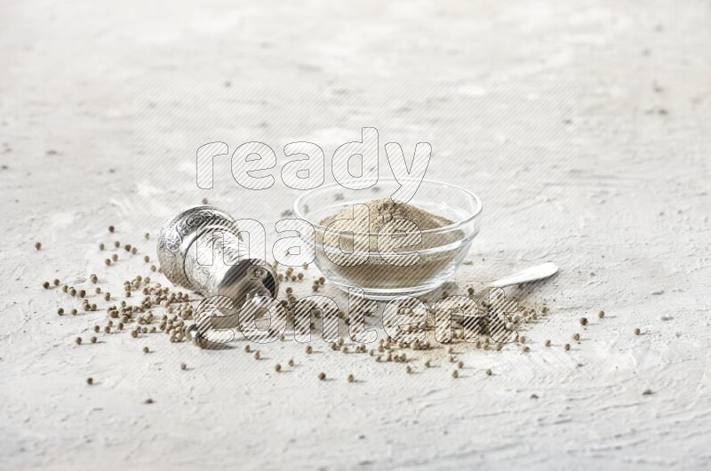 A glass bowl full of white pepper powder with white pepper beads and a metal grinder on textured white flooring
