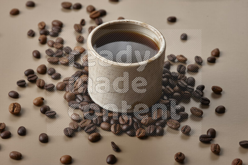 A beige pottery cup of coffee surrounded by roasted coffee beans on beige background