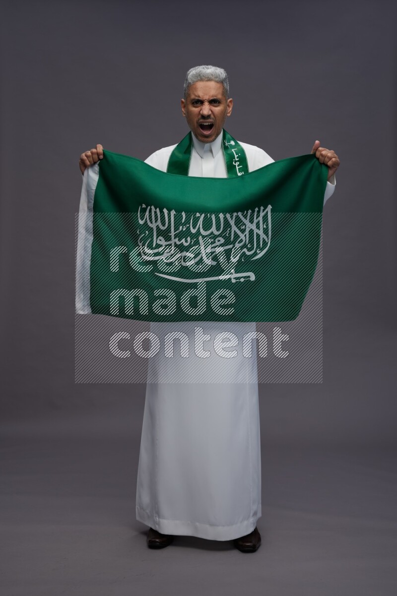 Saudi man wearing thob standing holding Saudi flag on gray background