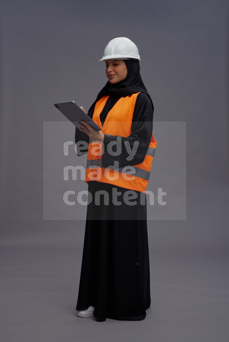 Saudi woman wearing Abaya with engineer vest standing working on tablet on gray background