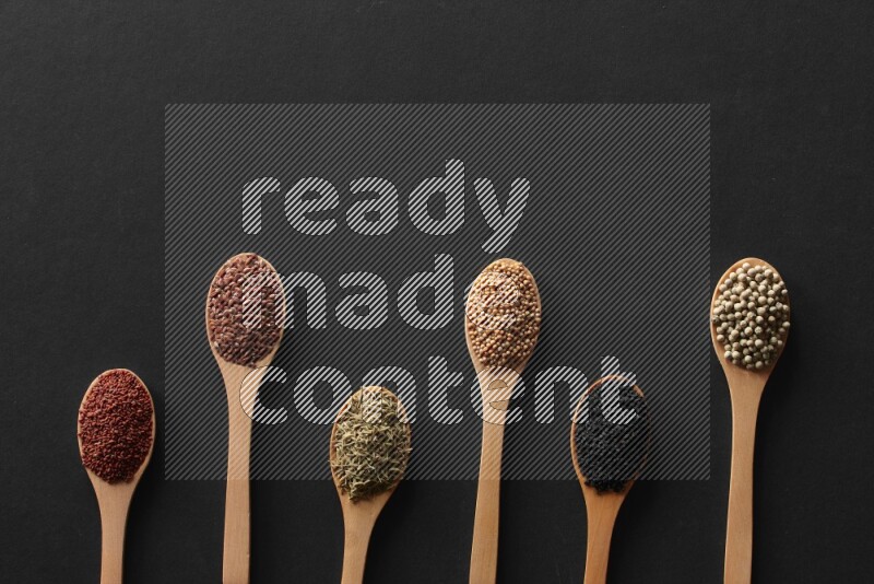 wooden spoons filled with white peppers, mustard seeds, black seeds, garden cress, cumin and flax on black flooring
