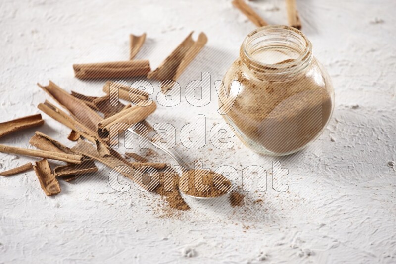 Herbal glass jar full cinnamon powder and a metal spoon surrounded by cinnamon sticks on a white background