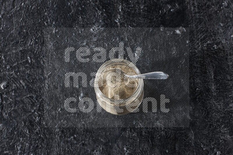 A glass jar and a metal spoon full of white pepper powder on textured black flooring