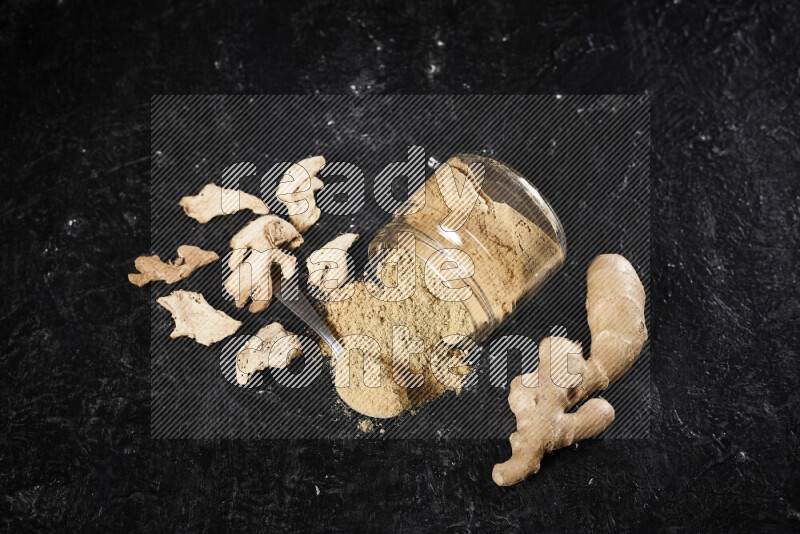 A glass jar full of ground ginger powder flipped with some spilling powder on black background