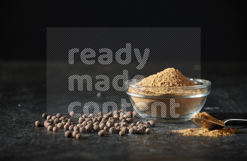 A glass bowl and metal spoon full of allspice powder and whole balls spreaded on a black flooring