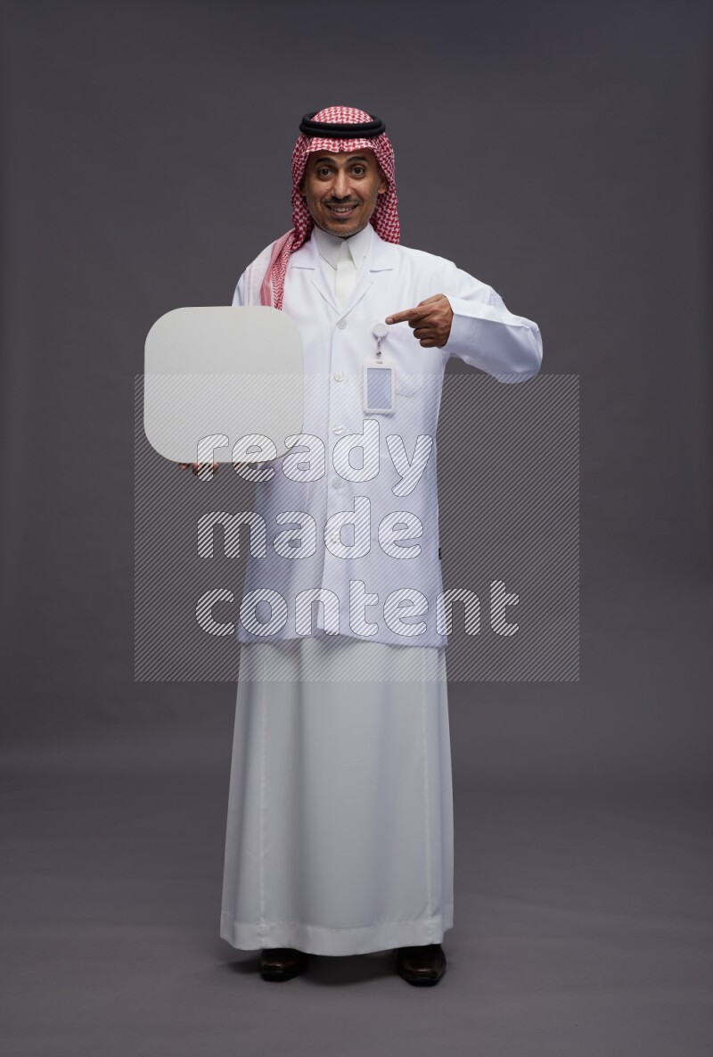Saudi man wearing thob with lab coat and shomag with pocket employee badge standing holding social media sign on gray background