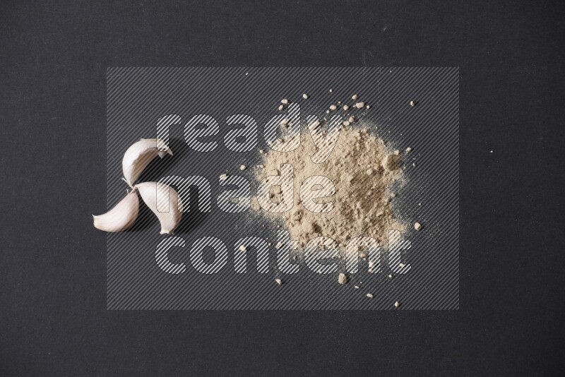 A small pile of garlic powder with some of cloves and a whole garlic bulb on a black flooring