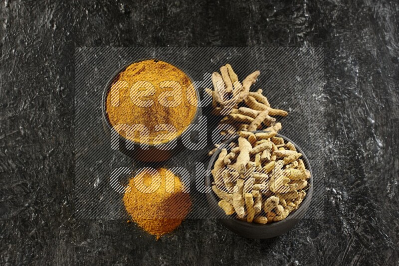 2 pottery black bowls, one full of turmeric powder and the other full of dried turmeric whole fingers on a textured black flooring