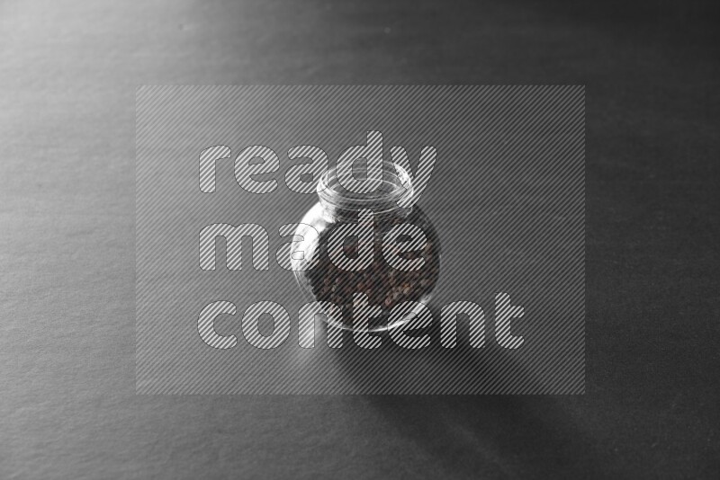 A glass spice jar full of black peppers on black flooring