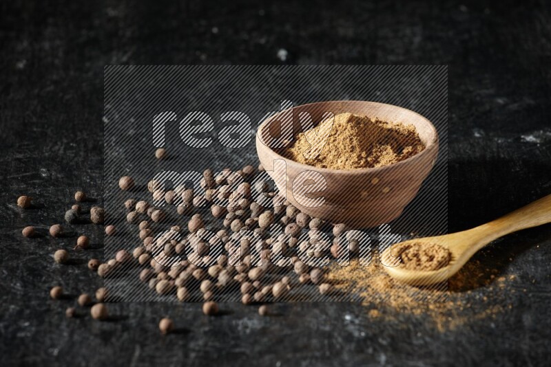A wooden bowl and spoon full of allspice powder and whole balls spreaded on a black flooring