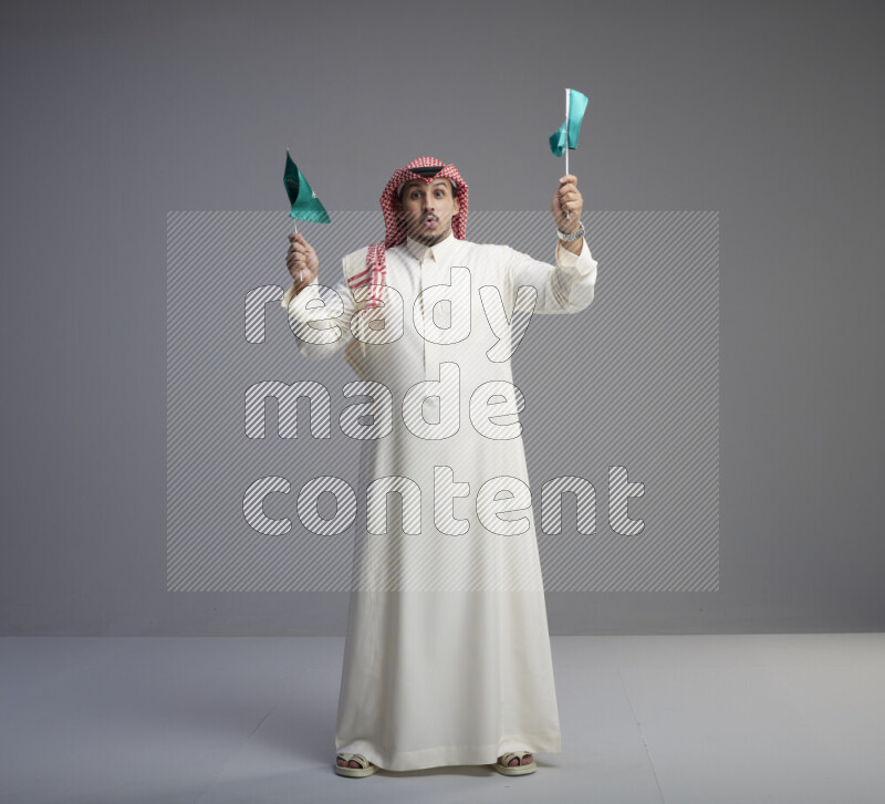A Saudi man standing wearing thob and red shomag raising small Saudi flag on gray background