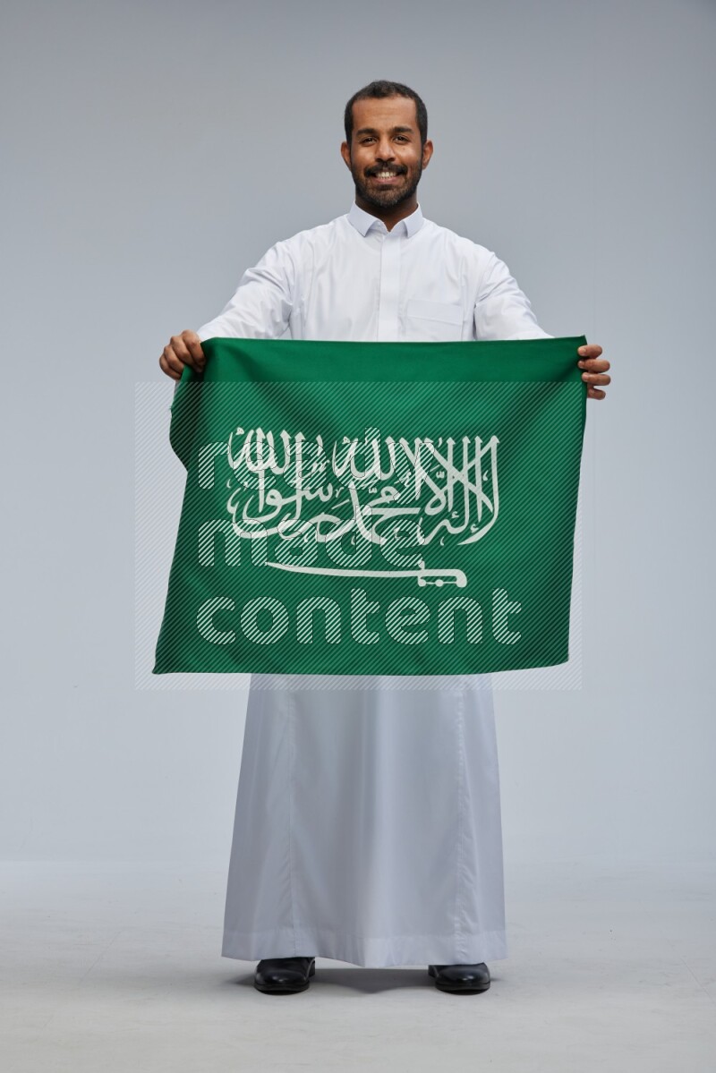 Saudi man wearing Thob standing holding Saudi flag on Gray background