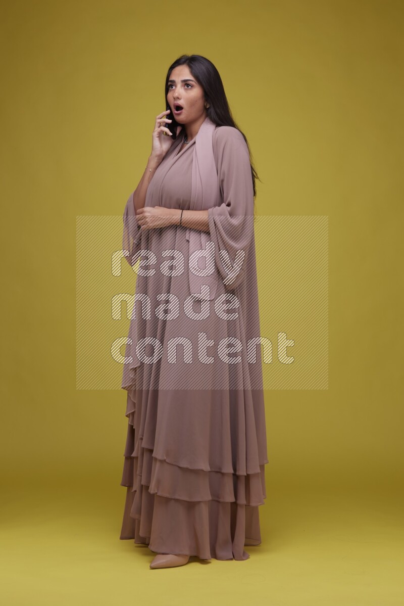 A woman Calling on a Yellow Background wearing Brown Abaya