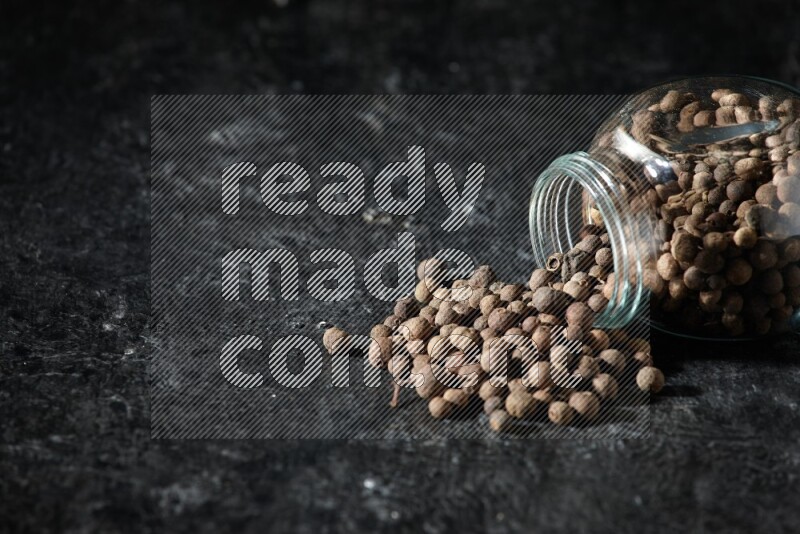 وعاء زجاجي للتوابل مقلوب ممتلئ بحبوب بهار الفلفل الحلو والحبوب تناثرت منه على خلفية سوداء