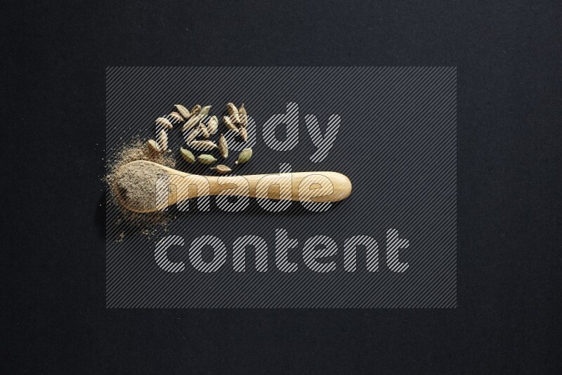 A wooden spoon full of cardamom powder and cardamom seeds beside it on black flooring