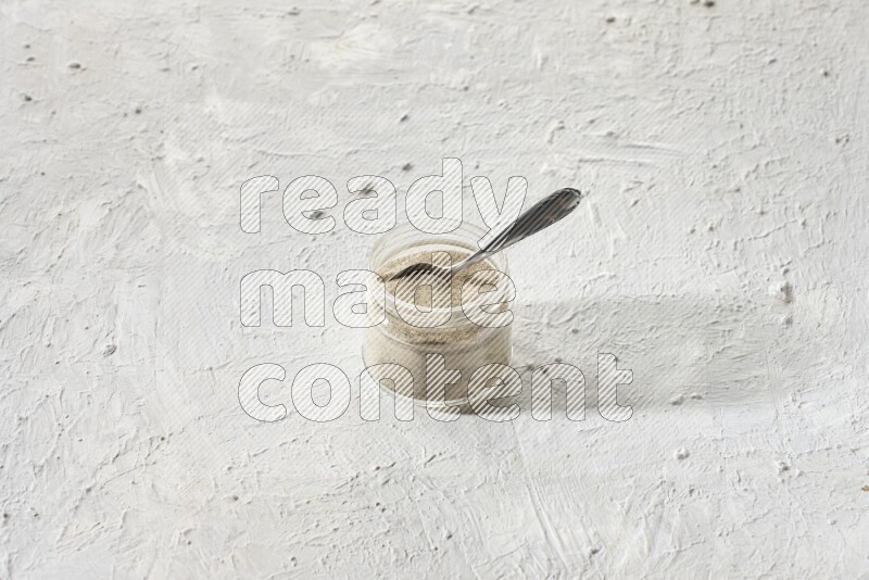 A glass jar and a metal spoon full of white pepper powder on textured white flooring