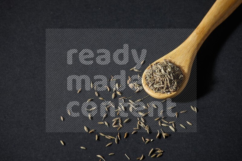 A wooden spoon full of cumin seeds on black flooring