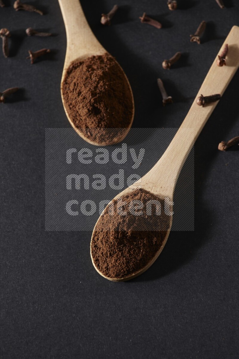 2 wooden spoons full of cloves powder with spreaded whole cloves on a black flooring