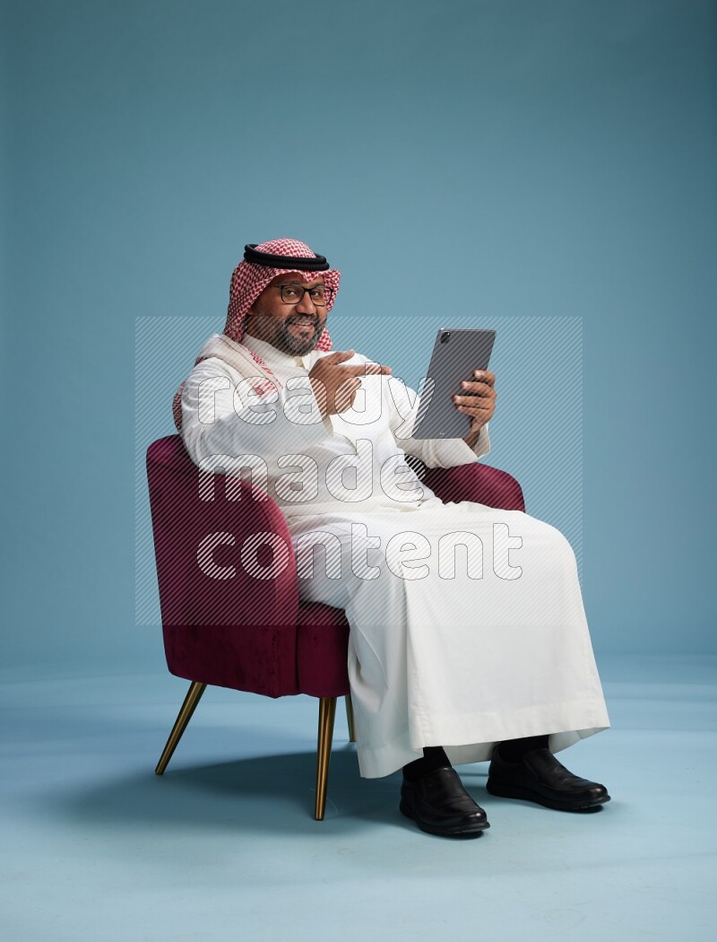 Saudi Man with shimag sitting on chair working on tablet on blue background